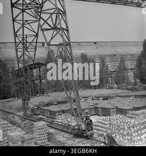 Sloviansk, 1981, AIZ (Armature-Insulator Plant). Ein genauerer Blick vom Boden aus mit Schwerpunkt auf die Schwermetallbasis und die Schienen eines massiven Portalkrans, der im Werkshof betrieben wird. Die Umgebung ist dicht gefüllt mit Stapeln von Keramikisolatoren und Holzkisten, die auf den Transport warten. Ein historisches Schwarzweiß-Archivfoto, das die authentische Atmosphäre der Epoche, die industrielle Logistikinfrastruktur und die Größenordnung der Fertigung festlegt. Ein detaillierter Einblick in die Vergangenheit, die sowjetische Industrie und den friedlichen Donbass. Stockfoto