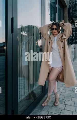 Trenchcoat für Damen, urbanes, stilvolles Modell, das in einem beigefarbenen Mantel und einer Sonnenbrille vor einem reflektierenden Glaseingangsbereich posiert Stockfoto