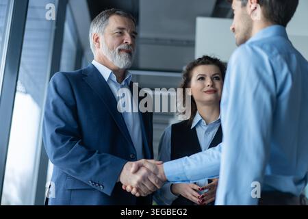 Glücklicher reifer Geschäftsmann, der sich mit einem jungen Geschäftsmann in einem modernen Büro die Hand schüttelt. Erfolgreicher Unternehmer begrüßt Geschäftsmann mit Handschlag. Vereinbarungen Stockfoto