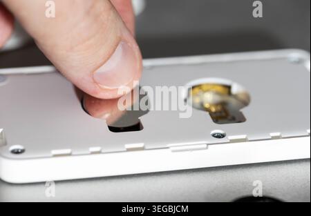 Detaillierte Ansicht der menschlichen Finger, die eine kleine Metallbatterie vorsichtig in den dafür vorgesehenen Schlitz des zerlegten weißen elektronischen Gadgets stecken. Aufgabenwiederholung Stockfoto