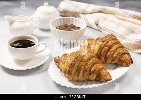 Frisch gebackene goldene Croissants werden auf einem weißen Teller mit einer Tasse schwarzen Kaffee und einer Schüssel Schokolade serviert, die typisch süße More symbolisiert Stockfoto