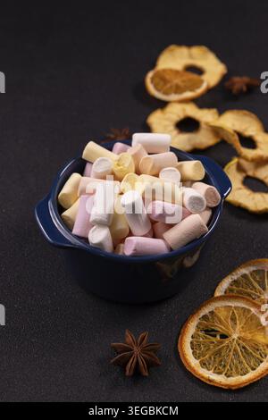 Bunte Marshmallows in einer blauen Schüssel, bereit für heiße Schokolade oder Kakao, umgeben von getrockneten Orangen, getrockneten Apfelscheiben und Sternanis auf einem dunklen Tex Stockfoto