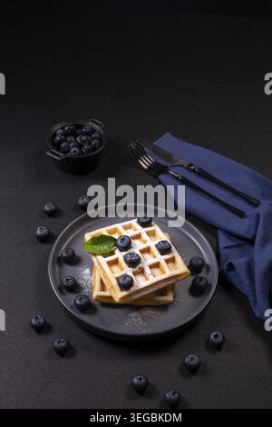 Stapel frisch gebackener Waffeln mit frischen Heidelbeeren, einer Prise Puderzucker und einem hellgrünen Minzblatt, das auf einem dunklen Teller liegt, zusammen mit Witz Stockfoto
