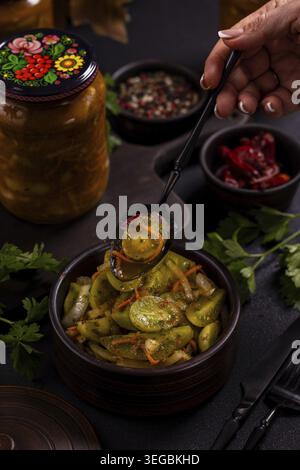 Servieren Sie selbstgemachte, eingelegte grüne Tomaten mit Karotten und Zwiebeln von Hand in einer rustikalen Schüssel neben einem dekorierten Glas mit Marmelade auf einer dunklen, gemütlichen Küche Stockfoto