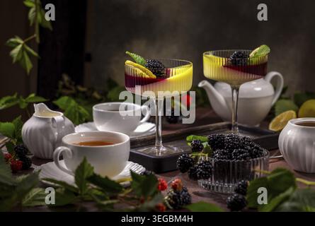 Zwei Gläser Panna Cotta-Dessert mit frischen Brombeeren, Zitronenscheiben und Minzblättern auf einem Tisch mit Teetassen, einer Teekanne und grünen Blättern t Stockfoto