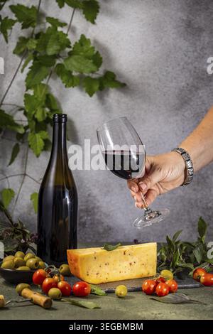 Halten Sie ein Glas Rotwein neben einer Flasche, Käse, Oliven und Kirschtomaten, um eine raffinierte kulinarische Szene zu schaffen Stockfoto