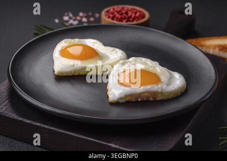 Essen, zwei herzförmige Spiegeleier auf einer schwarzen Keramikplatte auf dunklem Betonhintergrund. Frühstück zum valentinstag Stockfoto