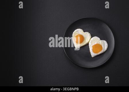 Essen, zwei herzförmige Spiegeleier auf einer schwarzen Keramikplatte auf dunklem Betonhintergrund. Frühstück zum valentinstag Stockfoto
