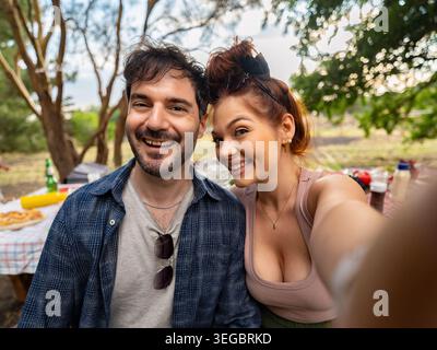Das fröhliche junge kaukasische Paar lächelt während eines Picknicks im Freien an einem Parktisch an einem sonnigen Sommertag, während es ein Selfie-Foto macht Stockfoto