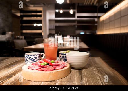 Rohe Wagyu-Rindfleischscheiben auf einem runden Holzbrett mit Chili-Pfeffergarnitur serviert, serviert zusammen mit einem rosa Limonaden und japanischen Keramikbechern, fotografiert in einem warm beleuchteten Tokio Yakiniku-Restaurant mit stimmungsvoller Bokeh-Einrichtung. Stockfoto
