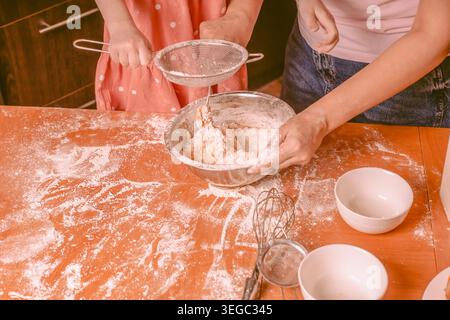 Ein Erwachsener und ein Kind sieben Mehl in eine Rührschüssel auf einem Holztisch, umgeben von Backutensilien und Mehl, das auf der Oberfläche verstreut ist Stockfoto