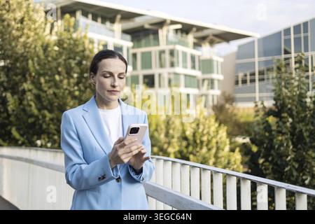 Geschäftsfrau steht draußen vor einem modernen, nachhaltigen Bürogebäude und blickt auf ihr Smartphone Stockfoto