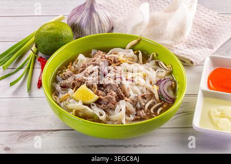 Eine leuchtend grüne Schüssel mit traditioneller vietnamesischer Rindfleischnudelsuppe Pho bo, mit Reisnudeln, Sojasprossen und Zwiebeln. Serviert mit frischer Limette, Chili, an Stockfoto