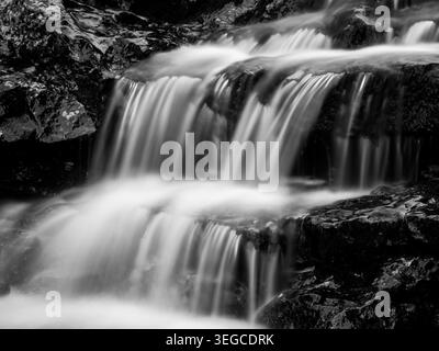 Lange Schwarzweiß-Exposition von gestuftem kaskadierendem Wasser über dunklen, nassen Felsen in einem felsigen Bach Stockfoto