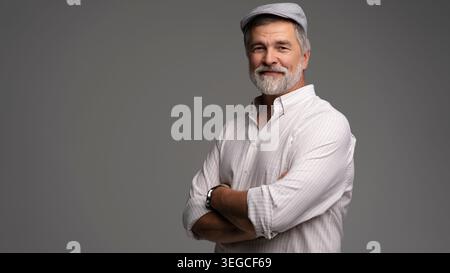 Lächelnder reifer Mann mit weißem Hemd und flacher Mütze, Nahaufnahme. Grauer Hintergrund Stockfoto