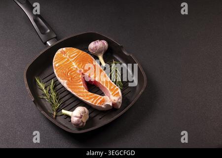 Rohes Lachssteak, frischer Knoblauch und grüne Rosmarinzweige in einer dunklen Grillpfanne, bereit für gesundes Kochen und die Zubereitung einer köstlichen, nahrhaften Mahlzeit Stockfoto
