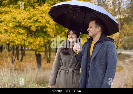 Glückliches Paar unter dem Schirm, das im Herbstregen im Park spaziert, mit Wärme und Liebe Stockfoto