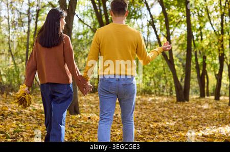 Ein junges Paar schlendert durch den Herbst Park und hält Hände zwischen sonnendurchfluteten Bäumen und Blättern Stockfoto