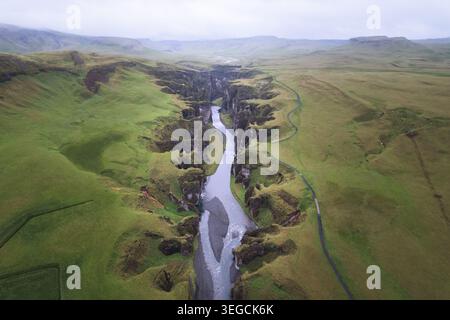 Aus der Vogelperspektive auf den Fjadrargljufur Canyon mit einem sich windenden Fluss, der zwischen steilen moosbedeckten Klippen unter einem bewölkten Himmel in Skaftarhreppur, Island fließt. Stockfoto