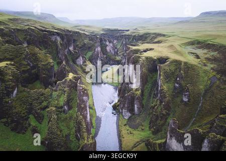 Aus der Vogelperspektive auf den Fjadrargljufur Canyon mit seinen steilen moosbedeckten Klippen und einem sich windenden Fluss unter dem bewölkten Himmel Skaftarhreppur, Island. Stockfoto