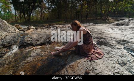 Frau, die auf Felsen sitzt und Füße in einem kleinen Wasserfall in einem sonnigen Wald tränkt Stockfoto