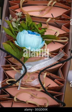 Schöne hellblaue künstliche Pfingstrose mit frischen grünen Blättern und silbernem Band, die auf ordentlich angeordneten rosa Geschenkboxen mit schwarzem Twist gelegt werden Stockfoto