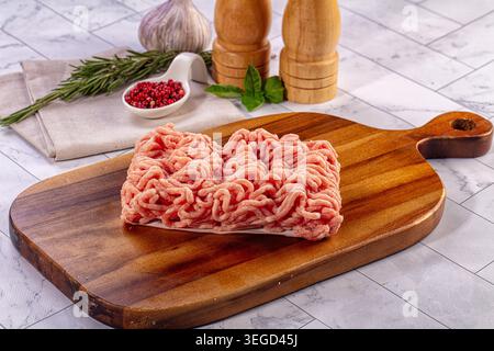 Frisch gemahlenes Geflügel auf Schneidebrett aus dunklem Holz. Kochszene mit Knoblauch, Rosmarin, Basilikum und rosafarbenen Pfefferkörnern. Hell beleuchtete Speisen c Stockfoto