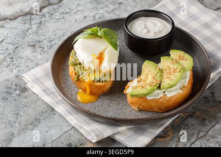 Nahaufnahme von zwei Toasts mit pochiertem Ei und Basilikumpesto, Avocado und Frischkäse serviert mit Sauerrahm auf einem Teller auf einem Tisch. Horizontal Stockfoto