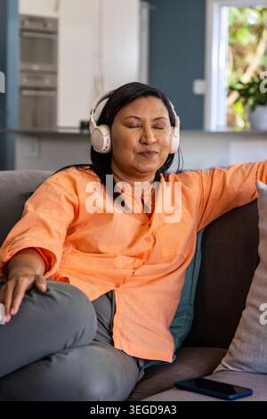 Reife asiatische Frau sitzt zu Hause auf grauem Sofa, hört auf weißen Kopfhörern, Smartphone daneben Stockfoto