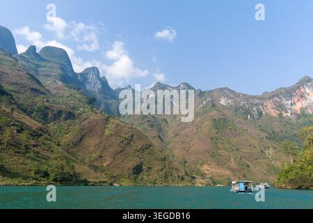Bergkette mit Blick auf den ruhigen türkisfarbenen See. Verstreute Wolken schweben im klaren blauen Himmel. Kleines Boot driftet in der Nähe der fernen Küste. Grünes und braunes Gemüse Stockfoto