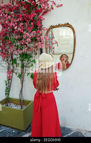 Modische Reisende mit Smartphone, die vor einem alten Spiegel und einer Blumenwand in der bezaubernden europäischen Altstadt posiert. Stockfoto