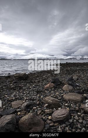 Blick auf eine felsige Küste entlang des Fjords mit schneebedeckten Bergen unter einem bewölkten Himmel in Tromso, Troms, Norwegen. Stockfoto