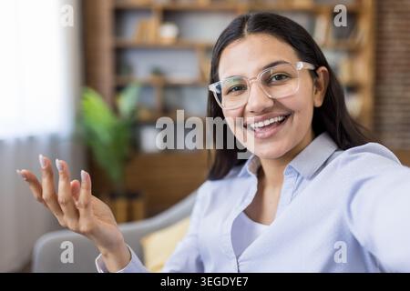 Junge Frau mit Brille kommuniziert gerne aus der Ferne oder fängt glückliche Momente ein, streckt die Hand, während sie direkt in die Kamera blickt, sitzt zuhause Stockfoto