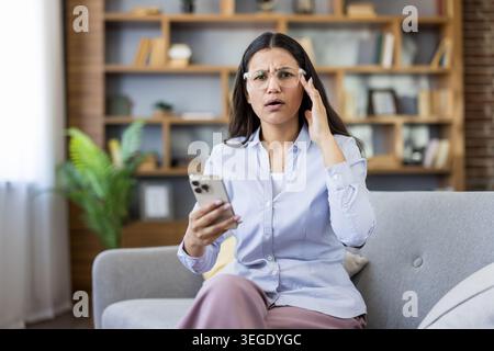 Eine junge Frau, die schockierende Nachricht auf ihrem Handy liest, mit einem verwirrten und besorgten Ausdruck, während sie ihren Kopf ungläubig hält, während sie zu Hause auf einer Couch sitzt Stockfoto