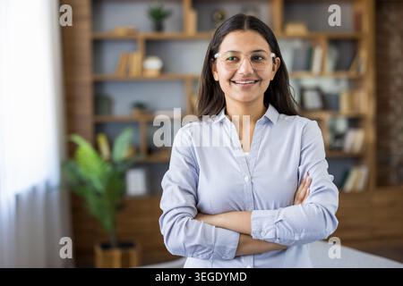 Selbstbewusste indische Geschäftsfrau lächelt und posiert mit gekreuzten Armen in einem modernen Büroumfeld, das beruflichen Erfolg, Führungsstärke und eine positive Einstellung repräsentiert Stockfoto
