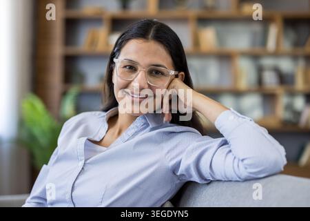 Eine junge Frau in einer klaren Brille und einem hellblauen Hemd lächelt selbstbewusst, während sie sich auf einem modernen Wohnzimmer-Sofa entspannt, gemütlich, zufrieden und mühelos stilvoll Stockfoto