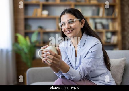Junge indianerin in Brille und lässiger Kleidung lächelt bequem auf einem Sofa, hält eine Kaffeetasse in einem gemütlichen, mit Büchern gesäumten Wohnzimmer und genießt eine friedliche Morgenpause Stockfoto