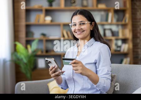 Lächelnde junge Erwachsene Frau, die eine Brille und ein blaues Hemd trägt, ein Mobiltelefon und eine Kreditkarte hält, Online-Shopping genießt und persönliche Finanzen von zu Hause aus verwaltet Stockfoto