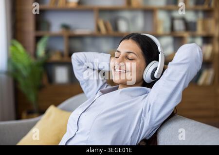 Die junge Frau im Kopfhörer lehnt sich mit geschlossenen Augen auf ein gemütliches Sofa zurück und genießt Musik und friedliche Entspannung zu Hause, lächelt und entspannt sich während ruhiger Auszeiten Stockfoto