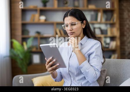Junge Frau in einer Brille auf einem Sofa, die ein Tablet hält, konzentriert und nachdenklich, während sie zu Hause recherchiert, Probleme löst und studiert, mit einem Bücherregal im Hintergrund Stockfoto