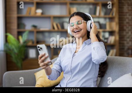 Junge Frau mit Kopfhörern und klaren Brillen, die einen fröhlichen Moment genießt, während sie Musik auf ihrem Smartphone hört, bequem auf einem Sofa in einer modernen Wohnumgebung sitzt Stockfoto