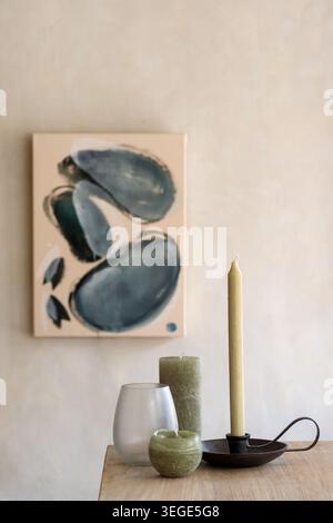 Eine Sammlung verschiedener dekorativer Kerzen, ein Glashalter und ein Metallkerzenhalter auf einem Holztisch mit einem abstrakten Leinwandgemälde im B Stockfoto