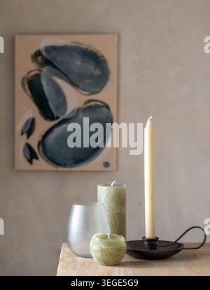 Eine Sammlung verschiedener dekorativer Kerzen, ein Glashalter und ein Metallkerzenhalter auf einem Holztisch mit einem abstrakten Leinwandgemälde im B Stockfoto