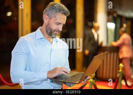 Geschäftsmann, der einen Laptop mit Nachtlicht benutzt. Geschäftsmann arbeitet auf der Straße am Laptop. Geschäftsmann mittleren Alters, der in der Nähe des Nachtclubs arbeitet. Business in Stockfoto