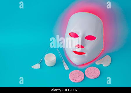 Trendige Hautpflege-Komposition mit futuristischer LED-Gesichtsmaske im Neonlicht, Gesichtscreme, Gua Sha, weißen Bürsten und Gesichtsschwämmen. Stockfoto