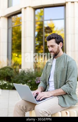 Mann, der außerhalb eines modernen Gebäudes an einem Laptop arbeitet und sich beim Tippen auf den Bildschirm konzentriert, repräsentiert flexiblen Lebensstil digitaler Nomaden und effiziente Produktivität in einer städtischen Umgebung Stockfoto
