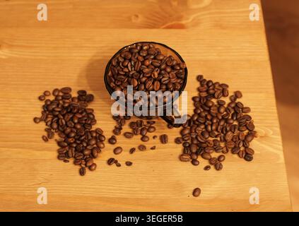Geröstete Kaffeebohnen, die aus einem überfüllten Messbecher auf einen hellen Holztisch gelangen, stehen für Energie, Aroma und den Beginn einer erfrischenden Tasse Stockfoto