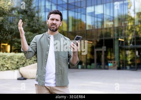 Mann draußen vor einem modernen Glasbürogebäude, der sein Smartphone mit frustriertem, verwirrtem Ausdruck und Gesten hält, während er mit schlechten Signalen und technischen Problemen zu kämpfen hat Stockfoto