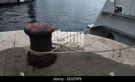 Nahaufnahme eines rostigen eisernen Schiffspollers mit einem Hochleistungs-Seeseil, das an einem Pier um ihn gebunden ist. Stockfoto