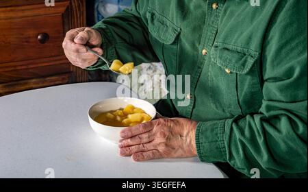 Eine Person sitzt an einem Tisch in einer Küche und hält einen Löffel mit Obst. Die Schüssel wird auf den Tisch gestellt. Das Licht ist weich, da es Nachmittag ist. Holzmöbel stehen im Hintergrund. Stockfoto